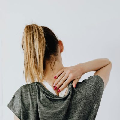 Back view of blond faceless lady in casual t shirt and manicure demonstrating pain spot on back while sitting near white wall